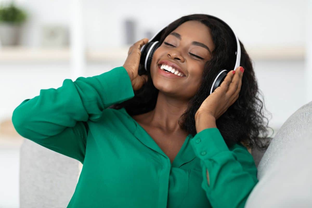 Une femme portant un chemisier vert sourit les yeux fermés tout en écoutant de la musique avec de gros écouteurs.