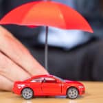 Une personne tient un petit parapluie rouge sur une voiture jouet rouge sur une surface en bois.