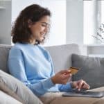 Une femme est assise sur un canapé, tenant une carte de crédit tout en utilisant un ordinateur portable, souriant et semblant effectuer un achat en ligne dans une pièce confortable et bien éclairée.