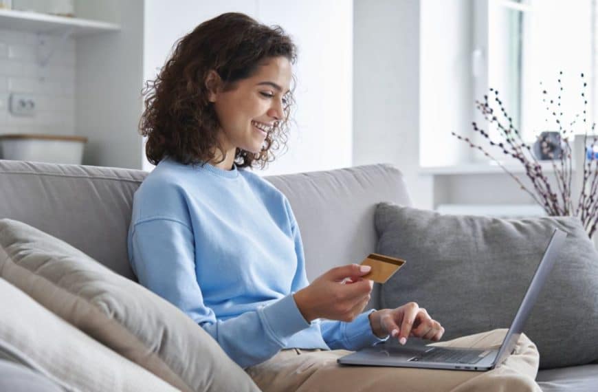 Une femme est assise sur un canapé, tenant une carte de crédit tout en utilisant un ordinateur portable, souriant et semblant effectuer un achat en ligne dans une pièce confortable et bien éclairée.