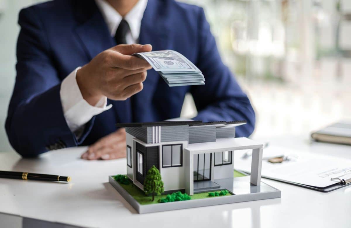 Une personne en costume remet une pile d'argent sur une table avec un modèle miniature d'une maison moderne dessus.