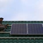 Une personne installe des panneaux solaires sur un toit de tuiles vertes sous un ciel nuageux.