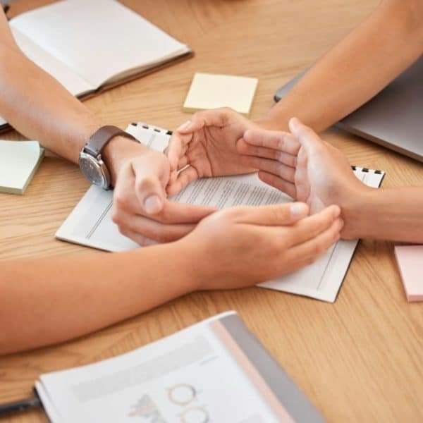 Quatre Personnes Sont Assises Autour D'une Table Avec Des Cahiers, Des Papiers Et Des Ordinateurs Portables, Tandis Que Deux D'entre Elles Se Tiennent La Main, Suggérant Un Travail D'équipe Ou Une Collaboration.