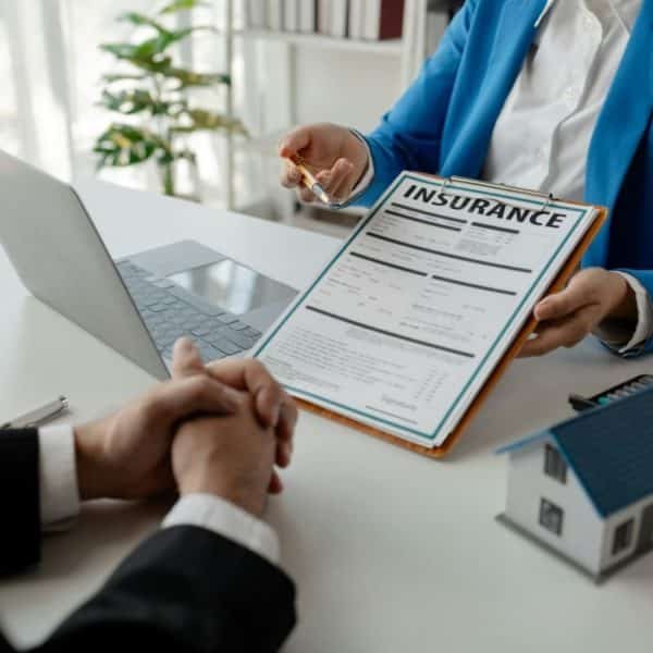 Deux Personnes Sont Assises à Un Bureau. L'une Tient Un Presse Papiers Avec Un Document D'assurance, Tandis Que L'autre écoute. Un Ordinateur Portable, Une Petite Maquette De Maison Et Une Calculatrice Sont Posés Sur La Table.