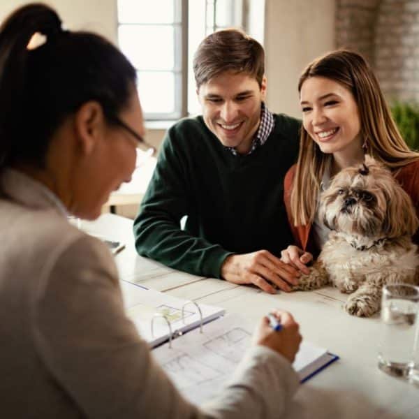 Un Couple Est Assis à Un Bureau Avec Un Chien, Interagissant Avec Une Femme Qui Tient Un Stylo Au Dessus D'un Dossier Ouvert.
