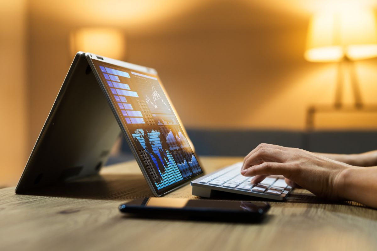 Une Personne Tape Sur Un Clavier Sans Fil Connecté à Un Ordinateur Portable En Mode Tente, Affichant Des Graphiques Colorés. Un Smartphone Est Posé à Proximité Sur Une Table En Bois.