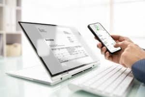 Une Personne Vérifie Une Facture Numérique Sur Un Smartphone Tandis Que Le Même Document Est Affiché Sur L'écran D'un Ordinateur Portable, Avec Un Clavier Blanc Et Une Souris à Proximité.
