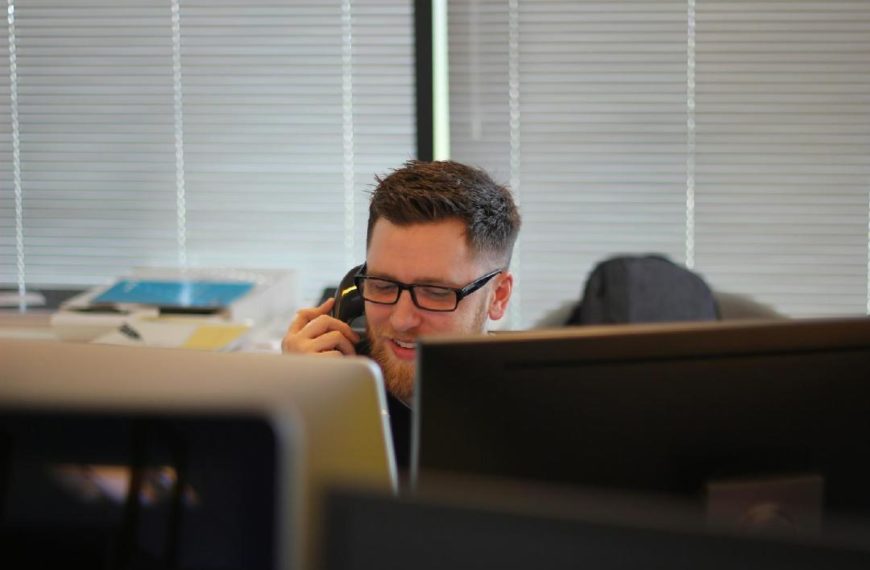 Un Homme Portant Des Lunettes Parle Au Téléphone Dans Un Bureau, Avec Des Ordinateurs Et Du Matériel De Bureau Autour De Lui.