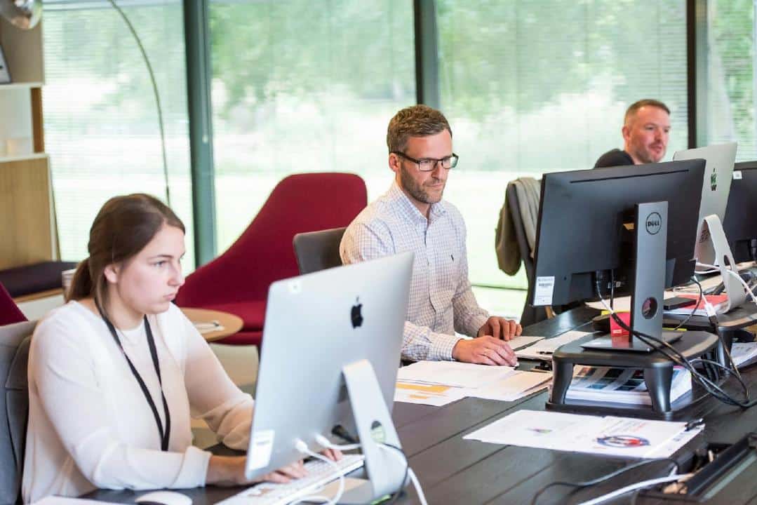 Trois Employés De Bureau Assis à Une Table Avec Des Ordinateurs, Concentrés Sur Leurs Tâches, Dans Une Pièce Avec De Grandes Fenêtres Montrant La Verdure à L'extérieur.