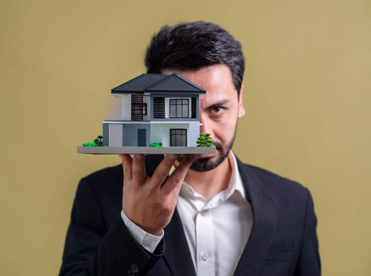 Un Homme En Costume Tient Une Petite Maquette De Maison Dans Sa Main Sur Un Fond Uni.