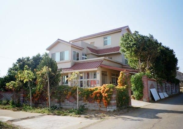 Maison à Deux étages Avec Un Toit De Tuiles Rouges, Entourée D'une Clôture Recouverte De Verdure Et De Fleurs Oranges, Située à L'intersection De Deux Routes Par Temps Clair.