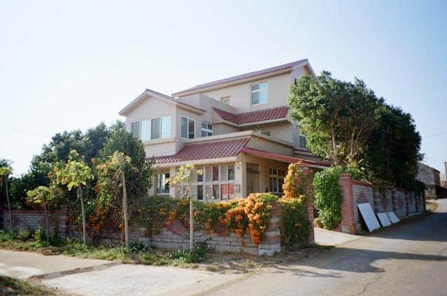 Maison à Deux étages Avec Un Toit De Tuiles Rouges, Entourée D'une Clôture Recouverte De Verdure Et De Fleurs Oranges, Située à L'intersection De Deux Routes Par Temps Clair.