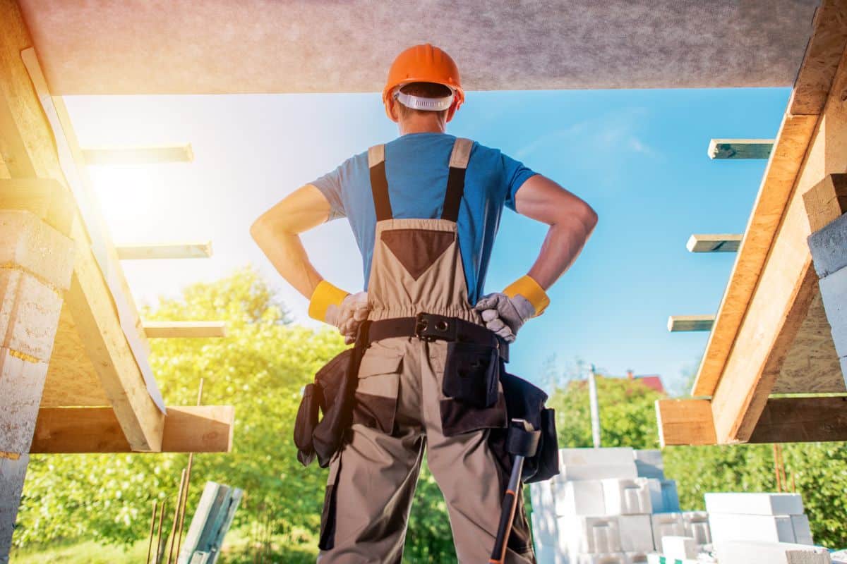 Un Ouvrier Du Bâtiment En Salopette Et Casque De Chantier Orange Se Tient Debout, Les Mains Sur Les Hanches, Observant Le Terrain De Qualité D'une Structure De Bâtiment Ouverte Sous Un Ciel Bleu Clair.