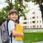 Une Fille Souriante Avec Des Nattes, Rayonnant D'assurance Scolaire, Tient Des Livres Et Porte Un Sac à Dos Tout En Se Tenant à L'extérieur Parmi Des Bâtiments Et Des Arbres.