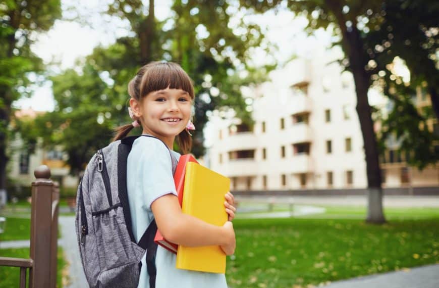 Une Fille Souriante Avec Des Nattes, Rayonnant D'assurance Scolaire, Tient Des Livres Et Porte Un Sac à Dos Tout En Se Tenant à L'extérieur Parmi Des Bâtiments Et Des Arbres.