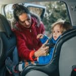 Un Adulte Portant Une Veste Rouge Attache Un Enfant Portant Une Veste Bleue Dans Un Siège Auto à L'intérieur Du Véhicule. Avec La Porte De La Voiture Ouverte, La Sécurité Est La Priorité.