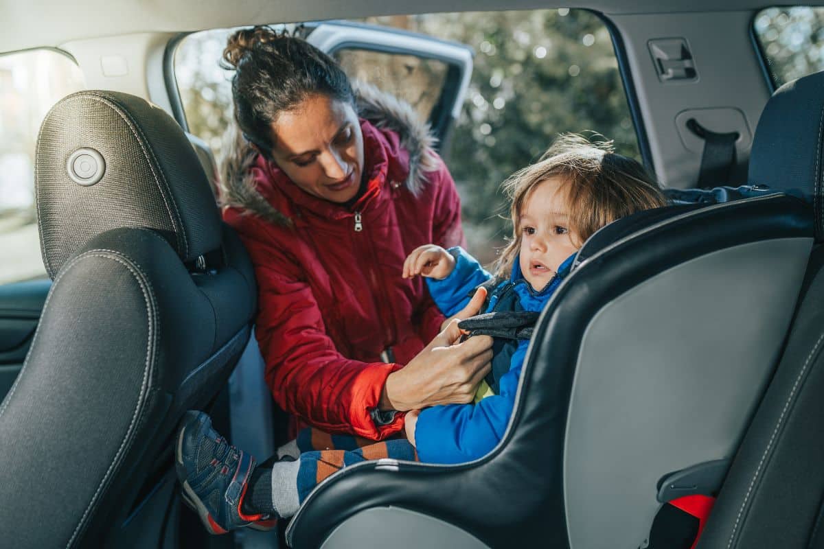 Un Adulte Portant Une Veste Rouge Attache Un Enfant Portant Une Veste Bleue Dans Un Siège Auto à L'intérieur Du Véhicule. Avec La Porte De La Voiture Ouverte, La Sécurité Est La Priorité.