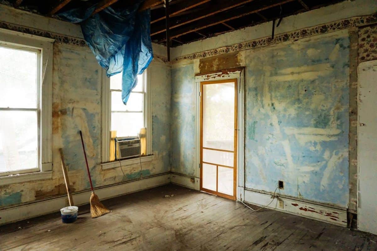 Ancienne Chambre En Cours De Rénovation, Poutres Apparentes Au Plafond, Murs Bleus écaillés, Deux Fenêtres, Un Climatiseur, Un Balai, Une Pelle à Poussière Et Une Bâche Bleue Suspendue Au Plafond.