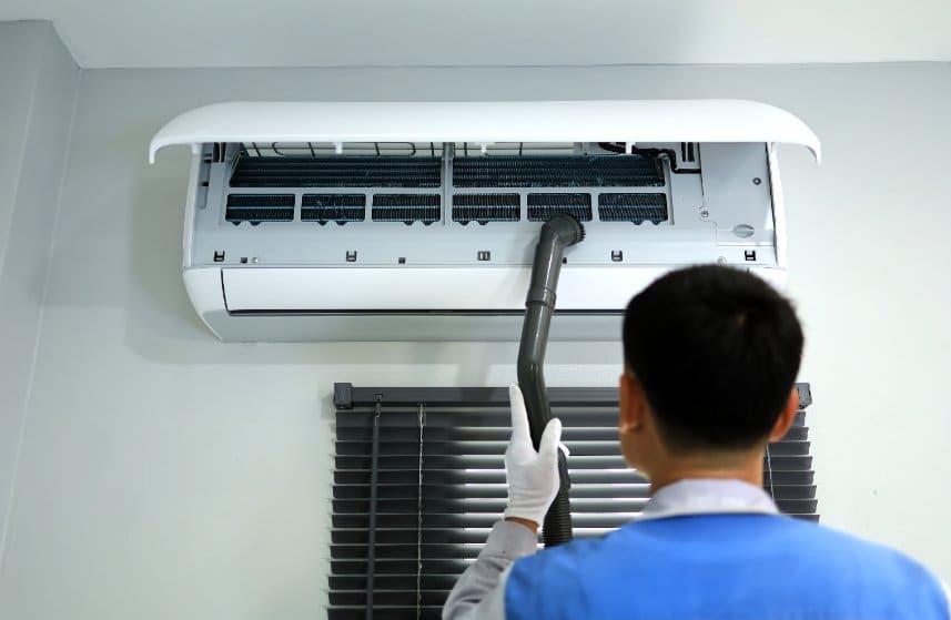 Technicien Nettoyant Un Climatiseur Avec Un Aspirateur, Debout Sur Une échelle Près D'une Fenêtre Aux Stores Fermés.