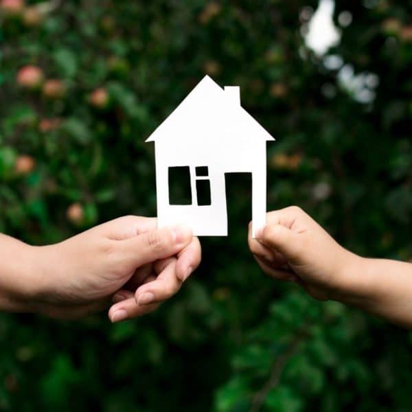 Deux mains tiennent une découpe de maison en papier blanc, symbolisant l'équilibre délicat de l'immobilier en zone protégée sur fond de feuillage vert luxuriant.