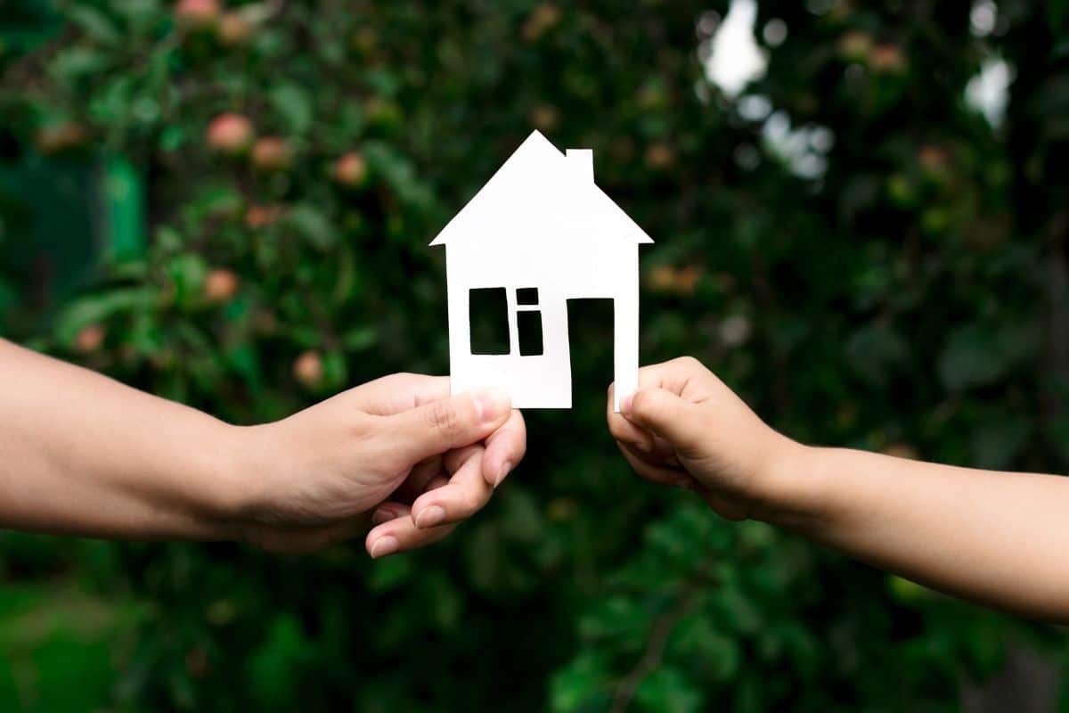 Deux mains tiennent une découpe de maison en papier blanc, symbolisant l'équilibre délicat de l'immobilier en zone protégée sur fond de feuillage vert luxuriant.