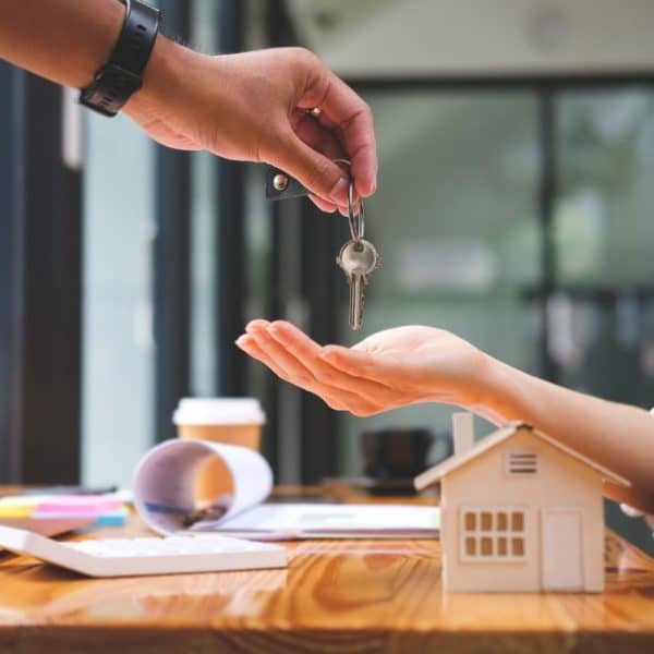 Une Personne Remet Les Clés à Une Autre, Dont La Main Est Ouverte, Symbolisant Un Nouveau Chapitre Dans Son Parcours De Financement Immobilier. Une Petite Maquette De Maison Et Des Fournitures De Bureau Reposent Sur La Table, Capturant L'essence Des Aspirations Immobilières.