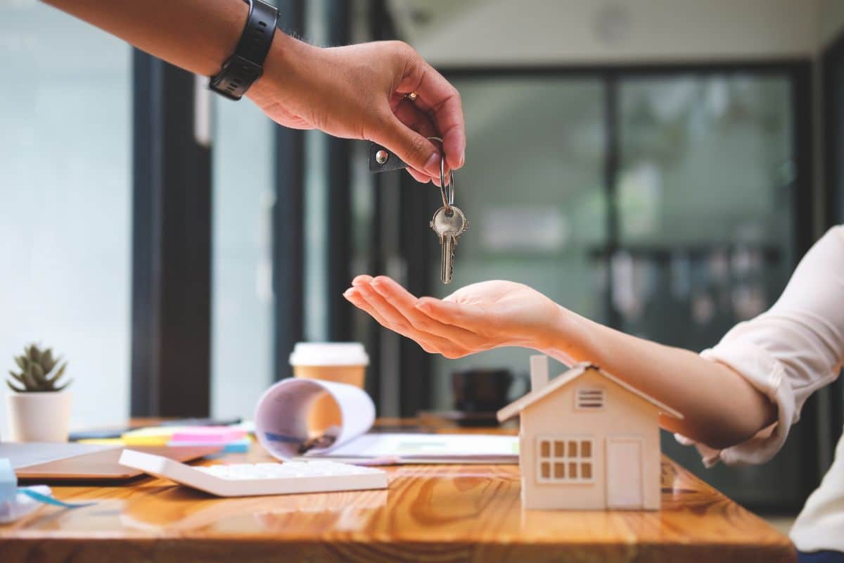 Une Personne Remet Les Clés à Une Autre, Dont La Main Est Ouverte, Symbolisant Un Nouveau Chapitre Dans Son Parcours De Financement Immobilier. Une Petite Maquette De Maison Et Des Fournitures De Bureau Reposent Sur La Table, Capturant L'essence Des Aspirations Immobilières.