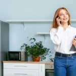 Une Femme En Chemise Blanche Parle Sur Un Smartphone D'« Immobilier à Distance » Tout En Tenant Un Presse Papiers, Debout Dans Une Cuisine Avec Une Plante En Pot, Un Micro Ondes Et Un Réfrigérateur.