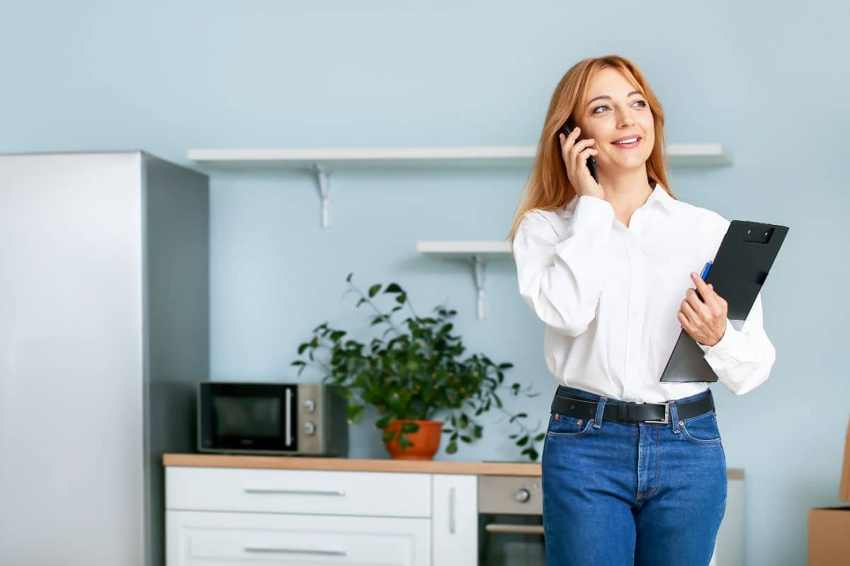 Une Femme En Chemise Blanche Parle Sur Un Smartphone D'« Immobilier à Distance » Tout En Tenant Un Presse Papiers, Debout Dans Une Cuisine Avec Une Plante En Pot, Un Micro Ondes Et Un Réfrigérateur.