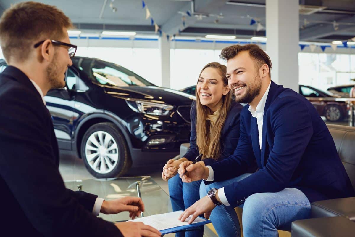 Un Couple Est Assis Chez Un Concessionnaire Automobile, écoutant Attentivement Un Vendeur Expliquer Les Avantages Du Kit De Conversion E8. Ils Semblent Engagés, Avec Des Voitures Garées Visibles En Arrière Plan.