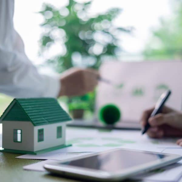 Deux Personnes Discutent De Documents Sur Une Table Ornée D'une Maquette De Maison, D'une Tablette Et D'une Plante En Pot. L'une Des Personnes Pointe Du Doigt Un Document Relatif à L'immobilier Durable Tandis Que L'autre Prend Des Notes Attentivement.