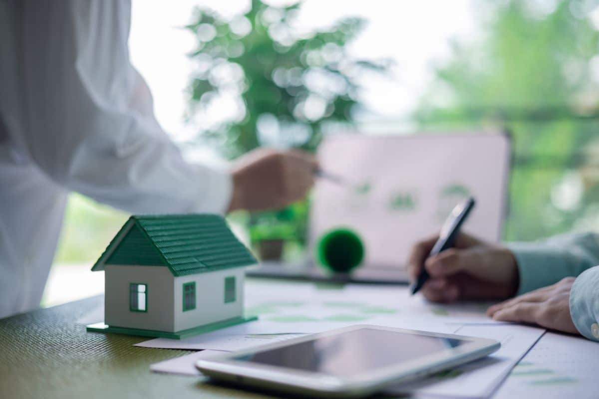 Deux Personnes Discutent De Documents Sur Une Table Ornée D'une Maquette De Maison, D'une Tablette Et D'une Plante En Pot. L'une Des Personnes Pointe Du Doigt Un Document Relatif à L'immobilier Durable Tandis Que L'autre Prend Des Notes Attentivement.