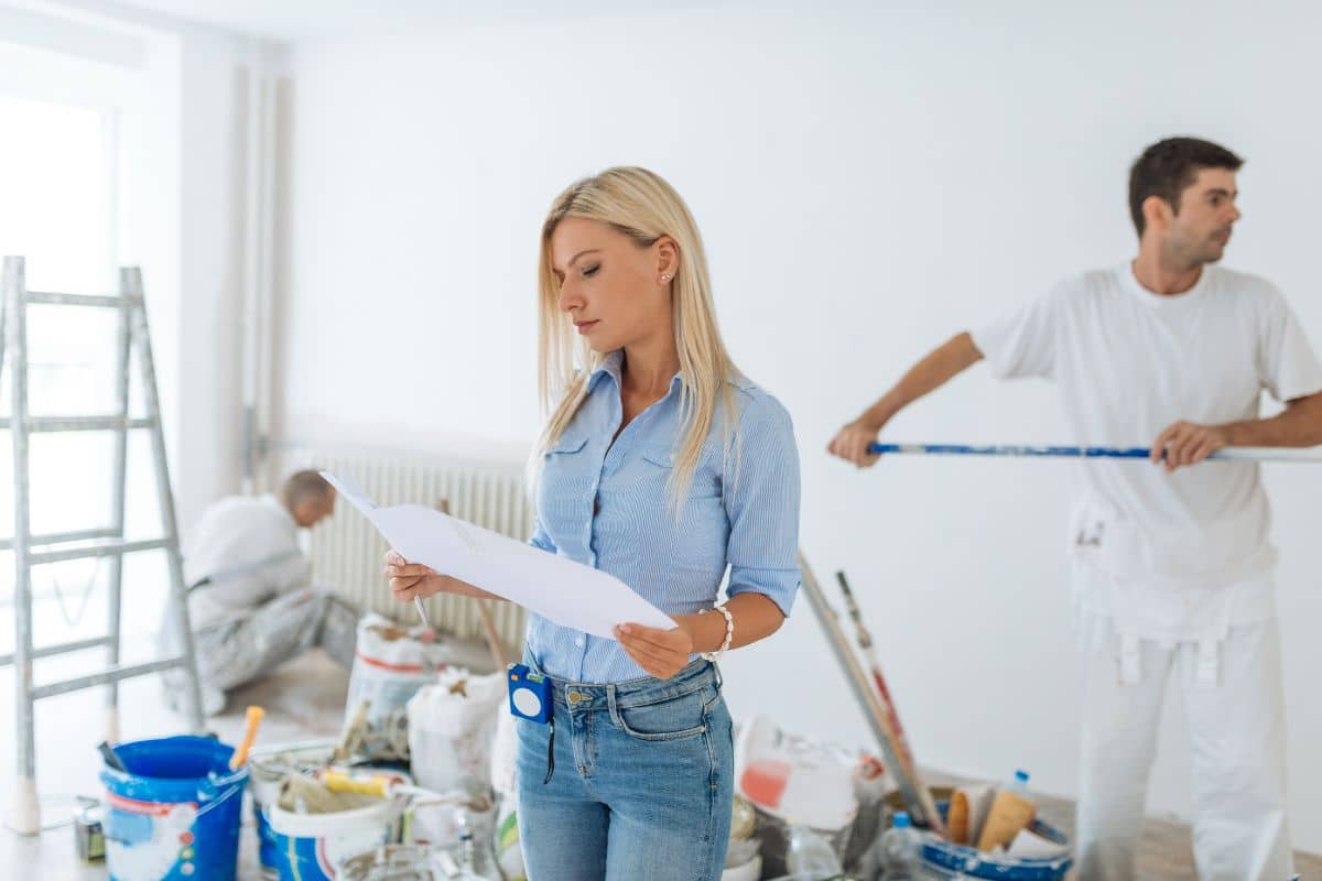 Une Femme En Chemise Bleue Examine Un Document Au Milieu De Travaux De Rénovation. Deux Hommes En Tenue De Travail Blanche Peignent ; L'un Est Sur Une échelle, Tandis Que L'autre Utilise Un Rouleau à Long Manche.