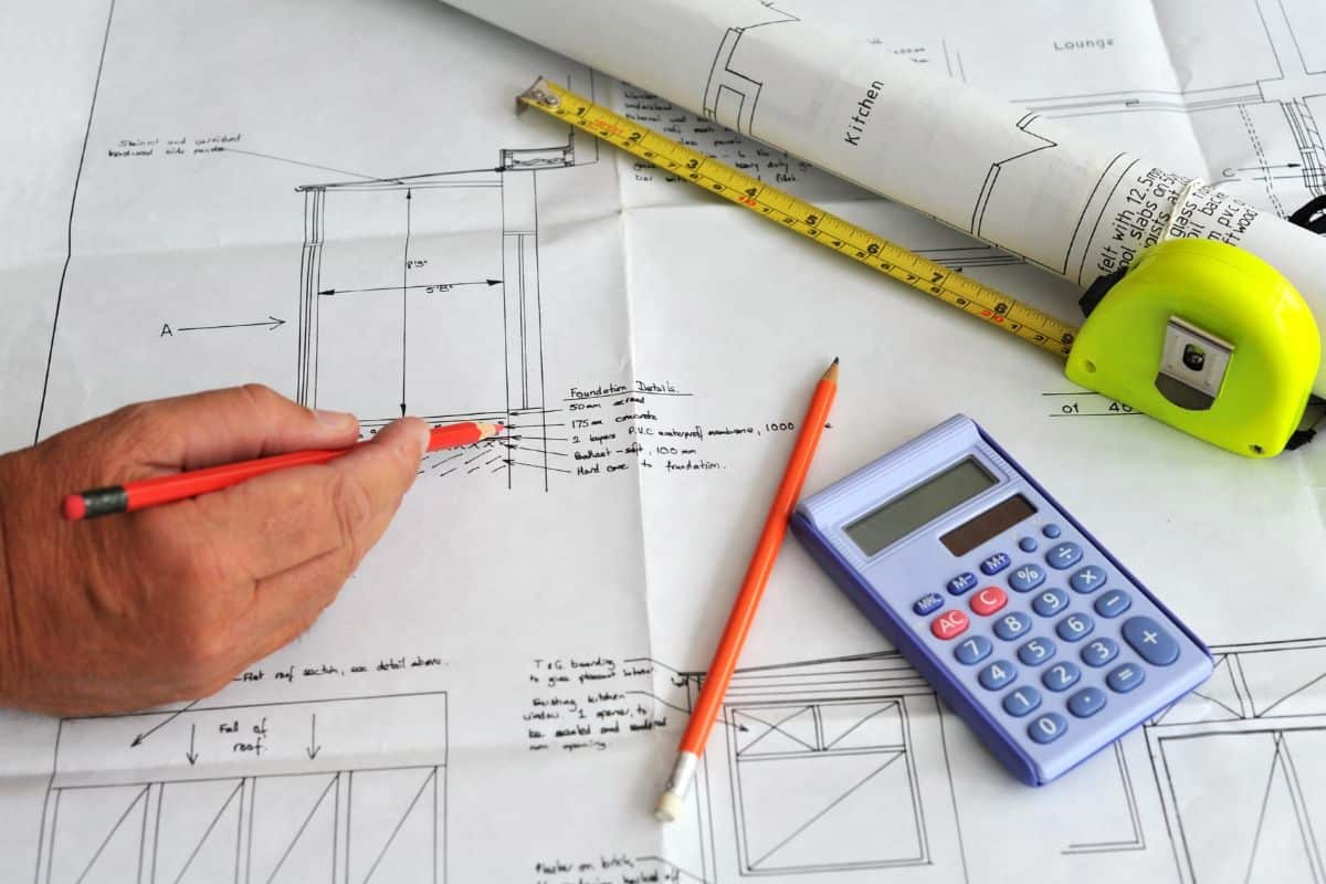 Dessin à La Main Sur Des Plans Architecturaux Avec Un Crayon Rouge, Planifiant Une Extension De Maison ; à Proximité Se Trouvent Une Calculatrice, Un Ruban à Mesurer Et Des Plans En Papier Roulé.