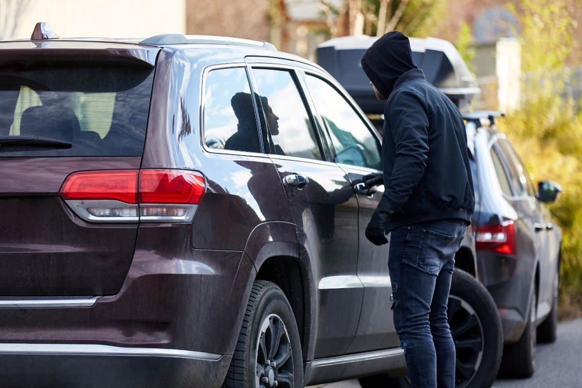 Une Personne Portant Un Sweat à Capuche Se Tient à Côté D'un Suv Sombre, Regardant à Travers La Fenêtre Côté Conducteur Du Véhicule à L'abri Du Vol étonnamment Sécurisé, Avec Une Autre Voiture Garée à Proximité.