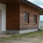 Maison Moderne Avec Revêtement De Façade En Bois, Avec Deux Fenêtres Et Une Porte De Garage. Des Guirlandes Lumineuses Ornent Le Toit, Projetant Une Lumière Chaleureuse. Au Premier Plan, Une Zone Herbeuse Luxuriante Complète Le Design épuré.