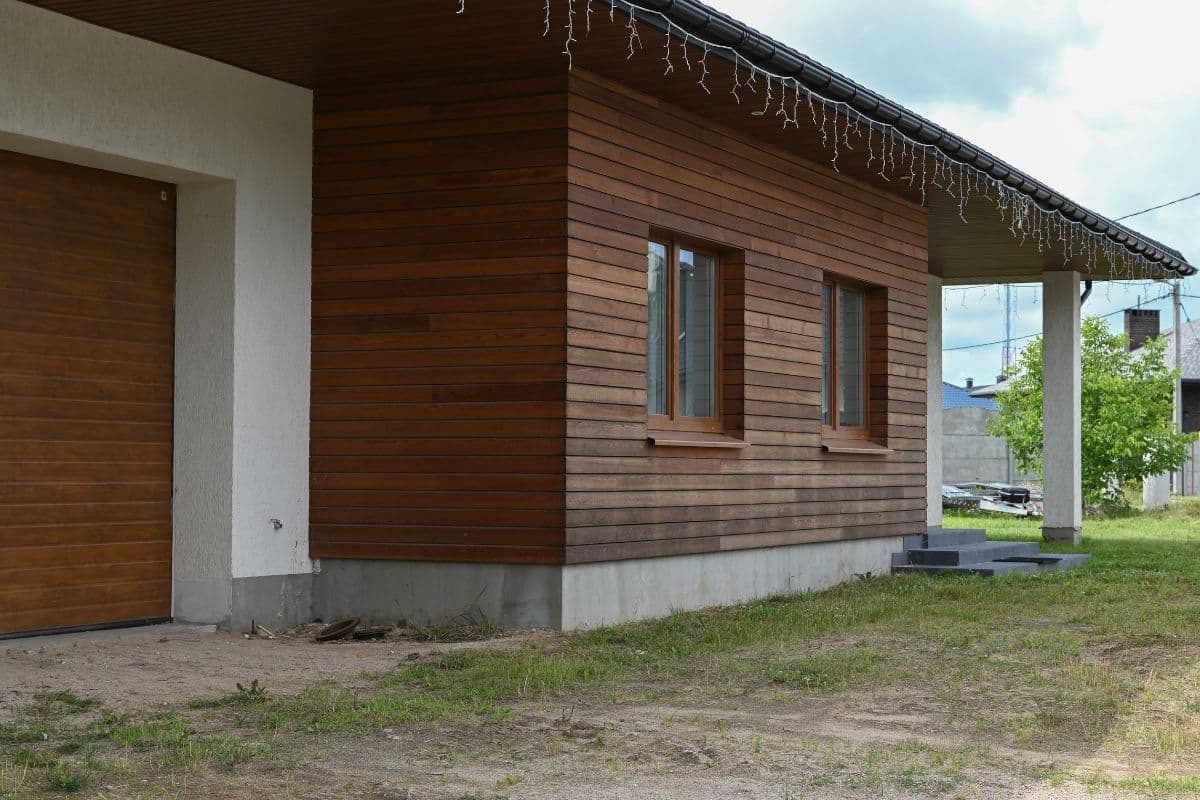 Maison Moderne Avec Revêtement De Façade En Bois, Avec Deux Fenêtres Et Une Porte De Garage. Des Guirlandes Lumineuses Ornent Le Toit, Projetant Une Lumière Chaleureuse. Au Premier Plan, Une Zone Herbeuse Luxuriante Complète Le Design épuré.