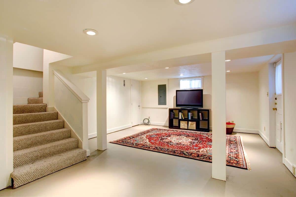 Une Pièce Au Sous Sol Aux Murs Beiges Et Au Tapis à Motifs Semble Transformée Par La Télévision Sur Pied Et L'escalier. Un éclairage Encastré Illumine L'espace.