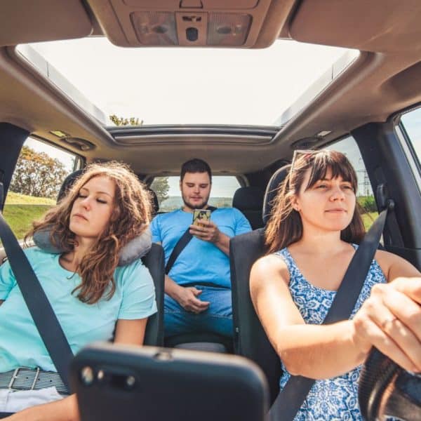 Trois Personnes Se Lancent Dans Un Long Voyage En Voiture ; Une Femme Conduit, Tandis Qu'un Homme Utilise Son Téléphone Sur Le Siège Arrière, Et Une Autre Femme Somnole Paisiblement Avec Un Oreiller De Nuque Sur Le Siège Passager.