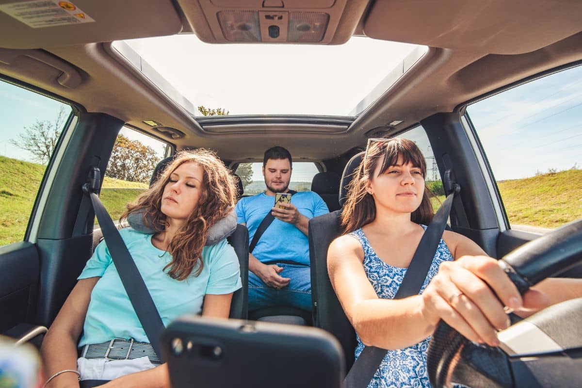 Trois Personnes Se Lancent Dans Un Long Voyage En Voiture ; Une Femme Conduit, Tandis Qu'un Homme Utilise Son Téléphone Sur Le Siège Arrière, Et Une Autre Femme Somnole Paisiblement Avec Un Oreiller De Nuque Sur Le Siège Passager.