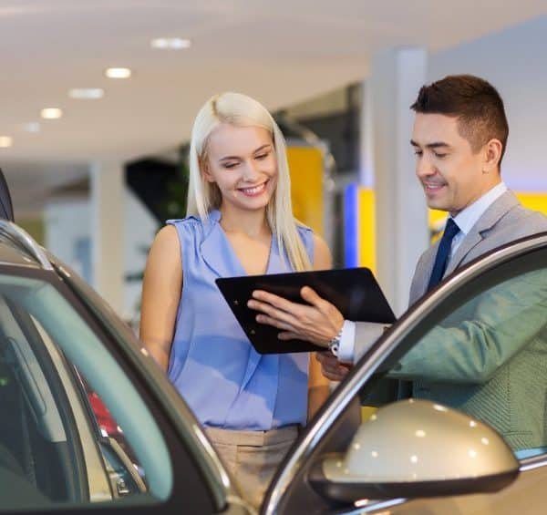Un Homme En Costume Montre Une Tablette à Une Femme à Côté D'une Portière De Voiture Ouverte à L'intérieur D'un Concessionnaire Automobile.