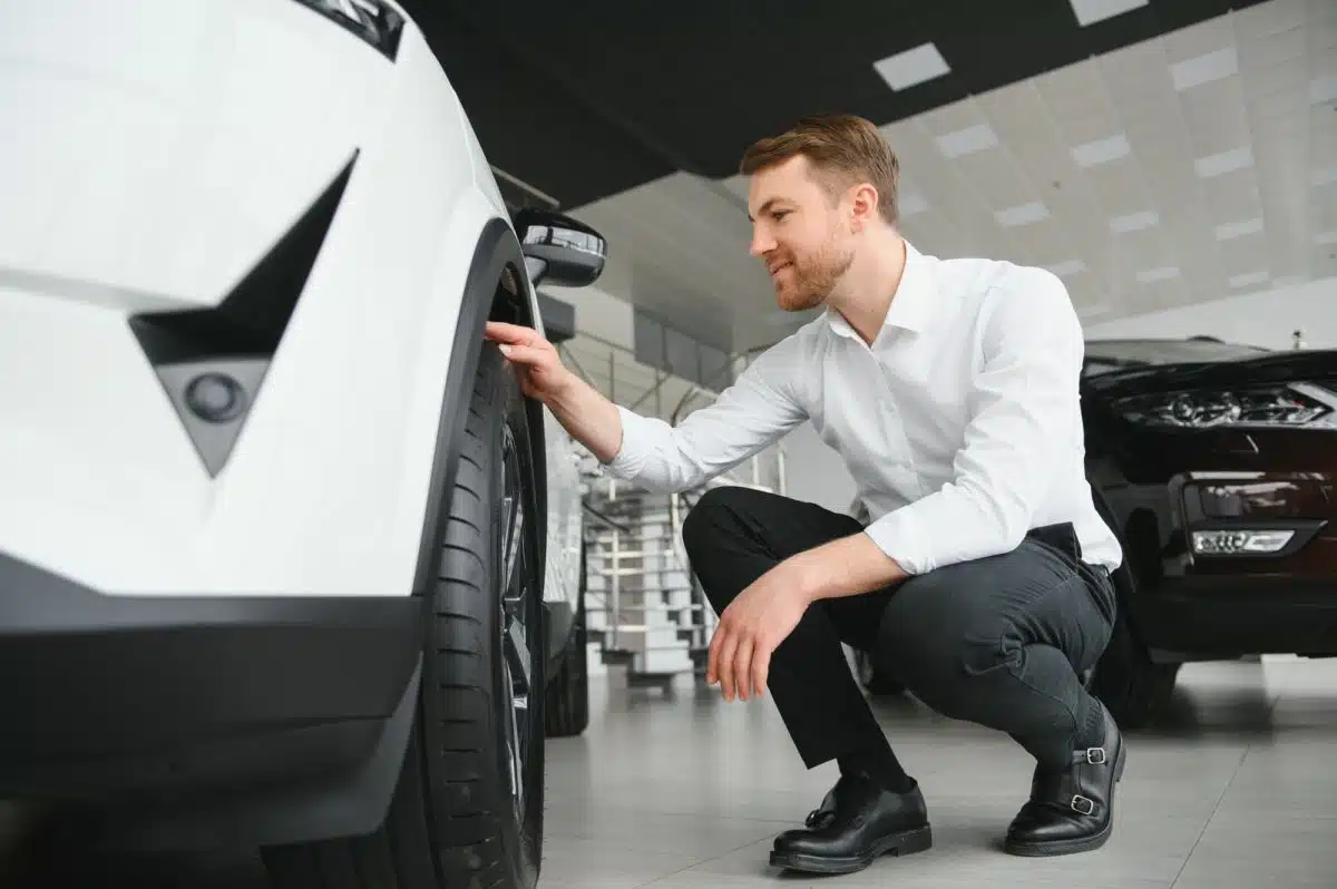 Un homme vêtu d'une chemise blanche et d'un pantalon foncé s'accroupit pour inspecter le pneu avant d'une voiture blanche à l'intérieur d'une salle d'exposition.