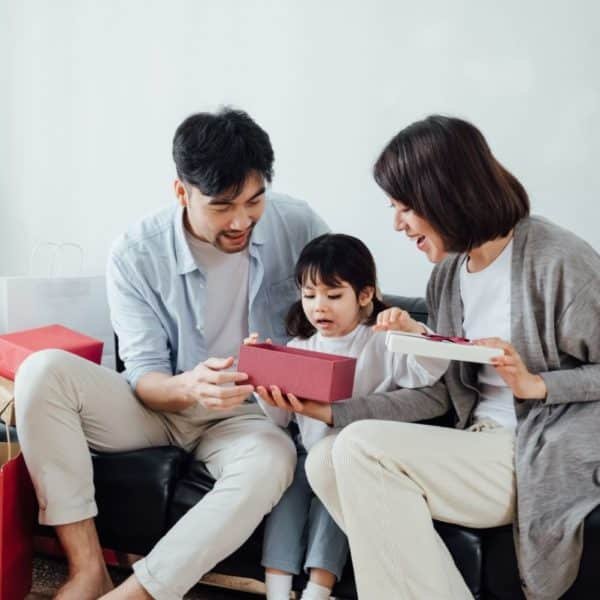 Une Famille De Trois Personnes Est Assise Sur Un Canapé, Ouvrant Ensemble Une Boîte Cadeau, Entourée De Sacs De Courses.