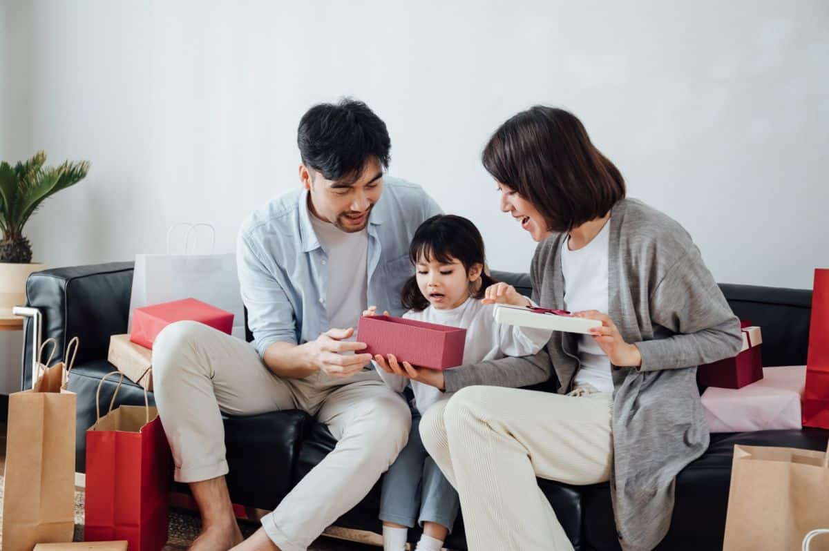 Une Famille De Trois Personnes Est Assise Sur Un Canapé, Ouvrant Ensemble Une Boîte Cadeau, Entourée De Sacs De Courses.