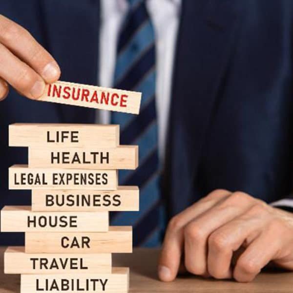 Une Personne En Costume Place Un Bloc De Bois étiqueté "assurance" Au Dessus D'une Pile De Blocs étiquetés Avec Des Catégories Telles Que Vie, Santé, Maison, Voiture, Voyage Et Responsabilité.