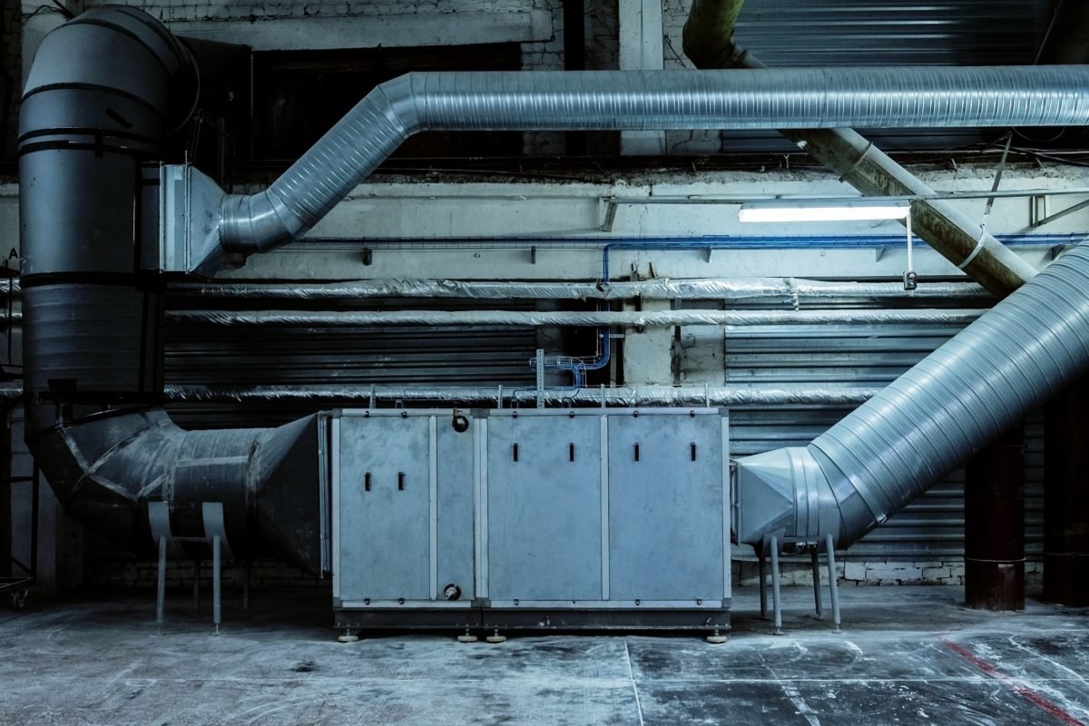 Dans Une Usine Ou Un Entrepôt, De Grands Conduits Et équipements De Ventilation Industrielle Sont Installés Le Long D'un Mur, Avec Une Tuyauterie Métallique Et Une Unité De Contrôle Visibles.