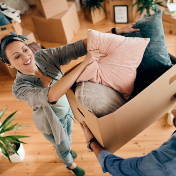 Deux Personnes Emballent Des Oreillers Dans Une Boîte En Carton Dans Une Pièce Au Sol En Bois, Entourée De Cartons De Déménagement Et D'une Plante En Pot.