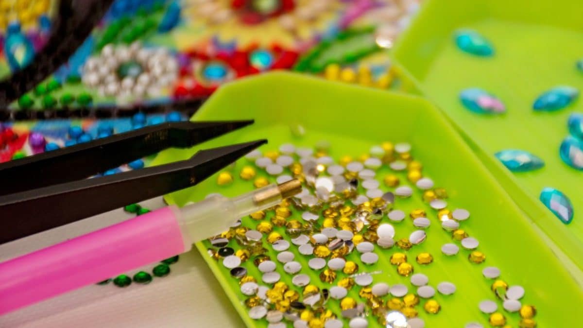 Gros Plan Sur Les Outils De Peinture Sur Diamant, Dont Un Stylet Rose, Une Pince à épiler Et Des Plateaux Verts Remplis De Petits Strass Colorés.