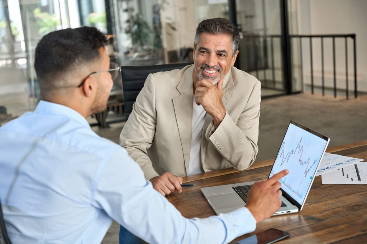 Deux Hommes Sont Assis à Un Bureau Et Discutent D'un Ordinateur Portable Affichant Des Graphiques Financiers. L'un D'eux Montre L'écran Tandis Que L'autre écoute En Souriant.