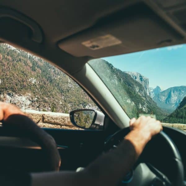 Une Personne Conduit Une Voiture Dans Un Paysage De Montagne Pittoresque, Visible à Travers Le Pare Brise, Avec Des Arbres Et Des Falaises Rocheuses Au Loin, Sous Un Ciel Dégagé.